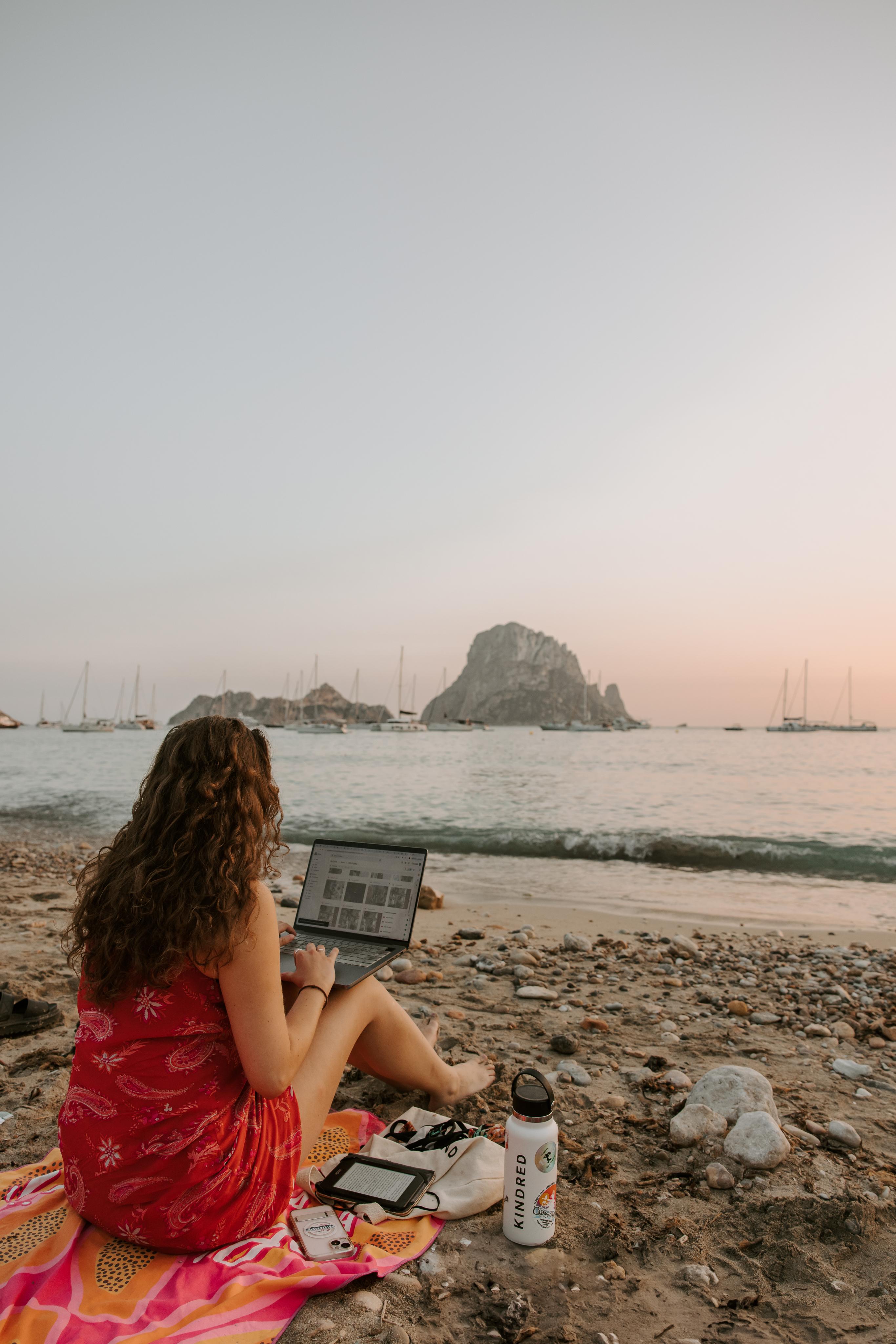 Une femme travaillant sur un ordinateur portable depuis une plage sereine au coucher du soleil, illustrant les possibilités de travail à distance avec l'échange de domiciles Kindred.