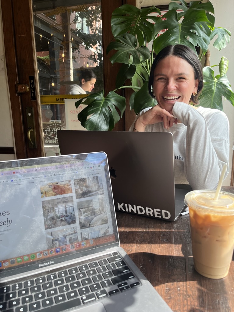 Smiling digital nomad working on a laptop at a cozy cafe, featuring a Kindred logo on the laptop. Perfect for remote work with a community vibe, flexible workspace, and great coffee.
