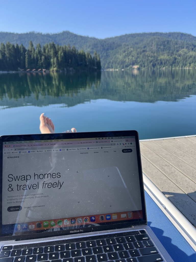 Lakeside remote work setup with a laptop showing the Kindred home exchange website, set against a backdrop of serene blue waters and lush green trees. Perfect for digital nomads seeking tranquility, nature, and flexible work environments.